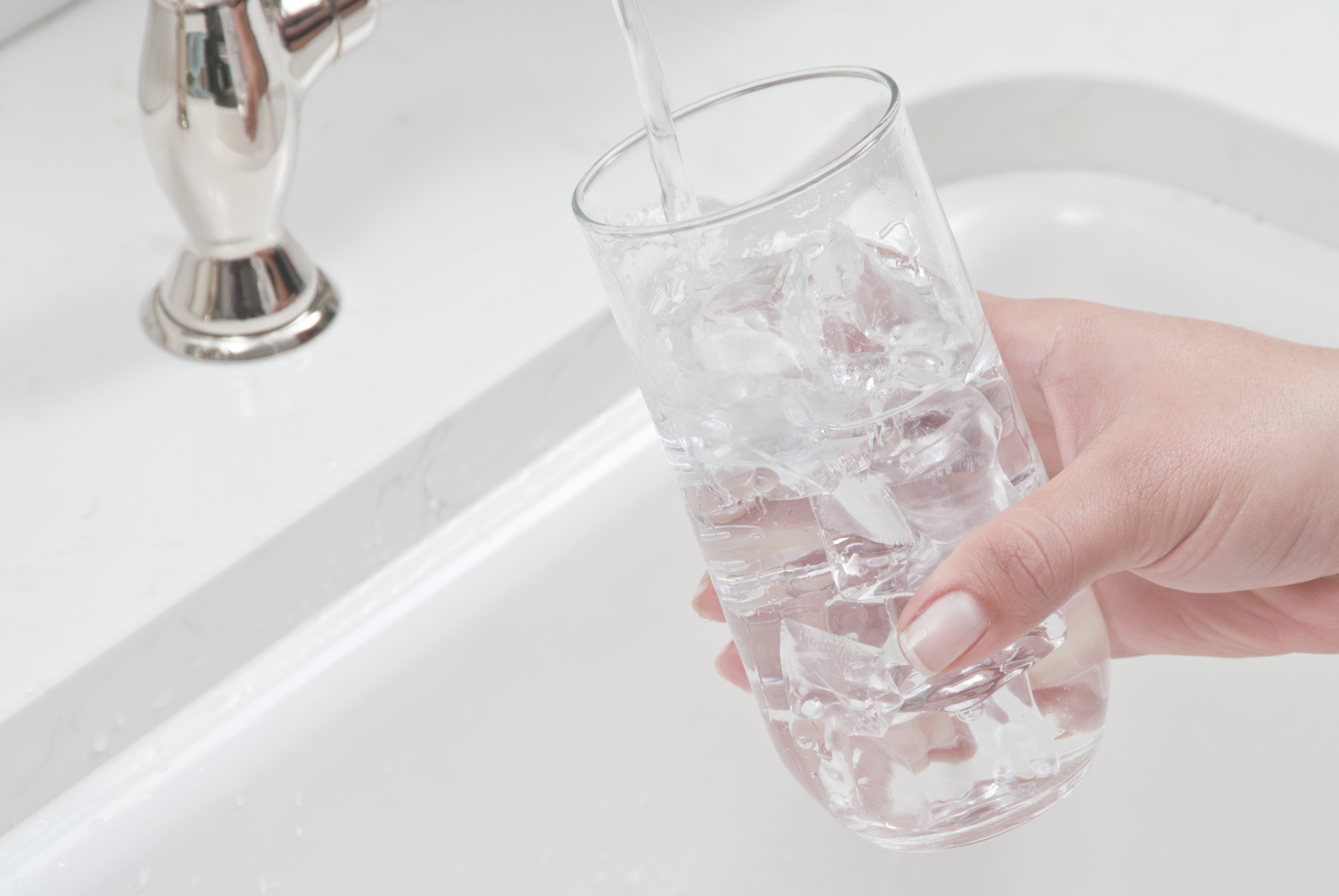 A hand holds a glass filled with ice under a running faucet, catching water in a white sink.
