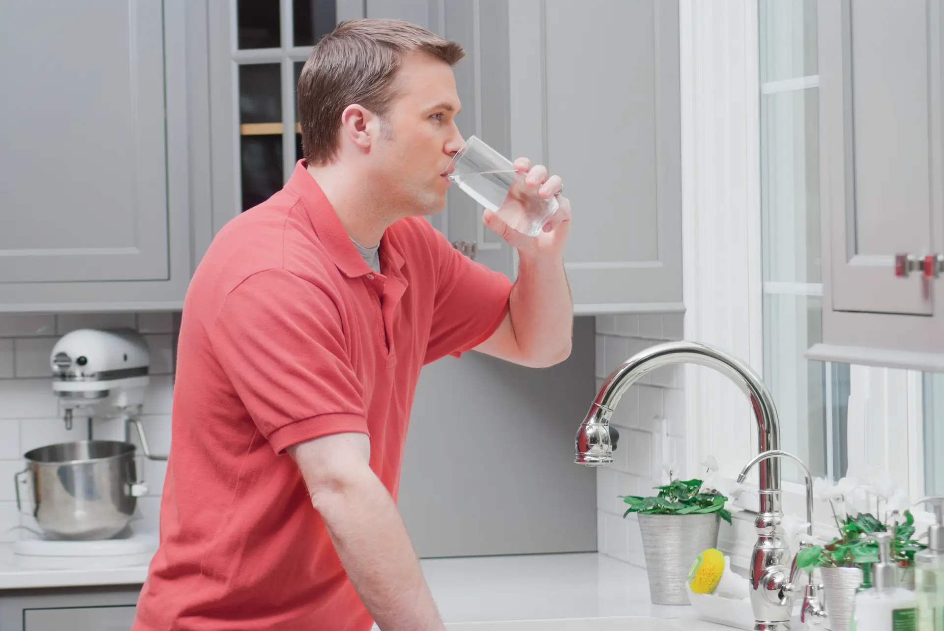 A man in a red polo shirt stands in a kitchen, drinking a glass of water by the sink. There are gray cabinets, a potted plant, and a mixer visible—highlighting the benefits of water conditioning in everyday life.
