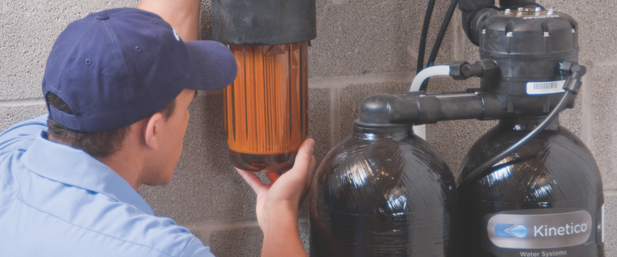 A technician in a blue uniform inspects a water treatment system mounted on a gray concrete wall, focusing on a clear filter housing with visible sediment.