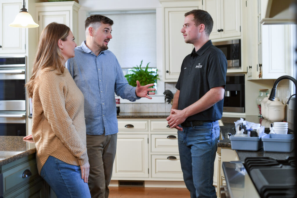 A man and woman stand in a bright, modern kitchen talking to a service professional in a black polo shirt about water softeners. They enjoy a friendly conversation near the counter as they discuss improving their home's water quality.