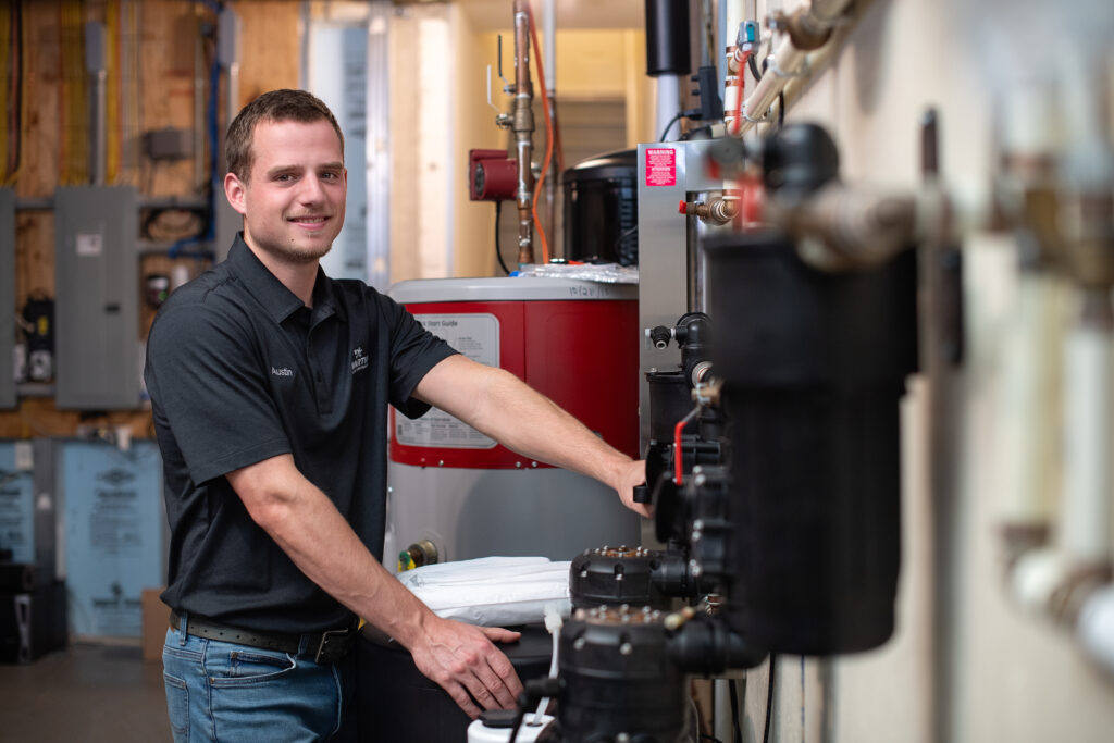 A man in a black polo shirt stands in a utility room, smiling while working on black water pumps and water softeners. Electrical panels and a water heater are visible in the background.