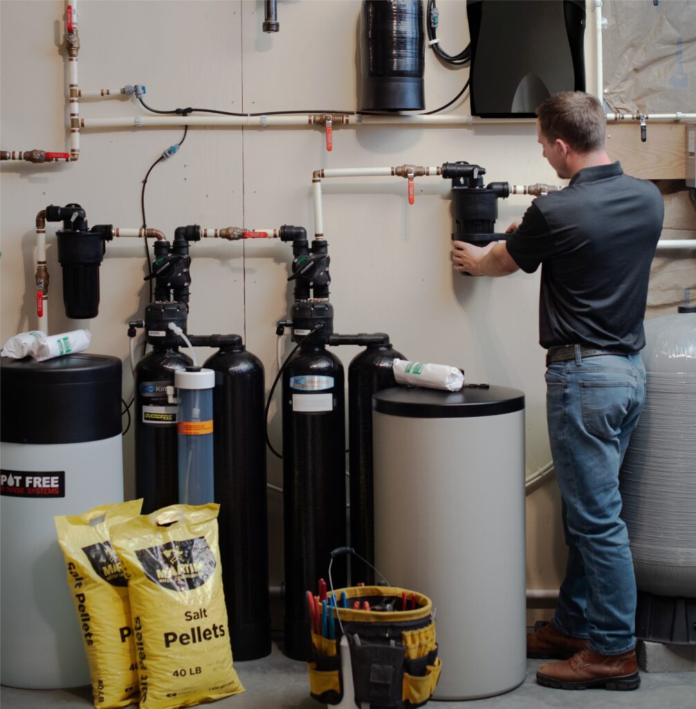 A man adjusts a water filtration system in a utility room. Multiple tanks, pipes, salt pellet bags, tools, water softener components, and other water treatment equipment are visible against the wall.