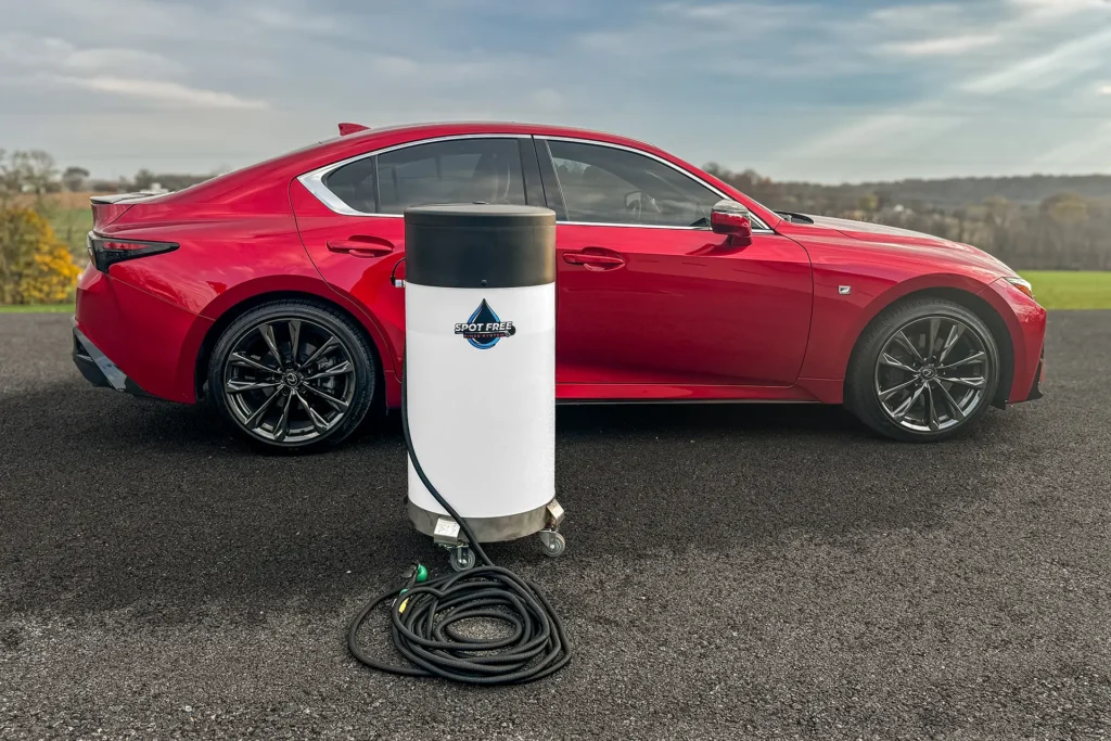A red car is parked on an asphalt surface outdoors with a white cylindrical device on wheels and a black hose positioned in front of it. The sky is partly cloudy and there is greenery in the background.