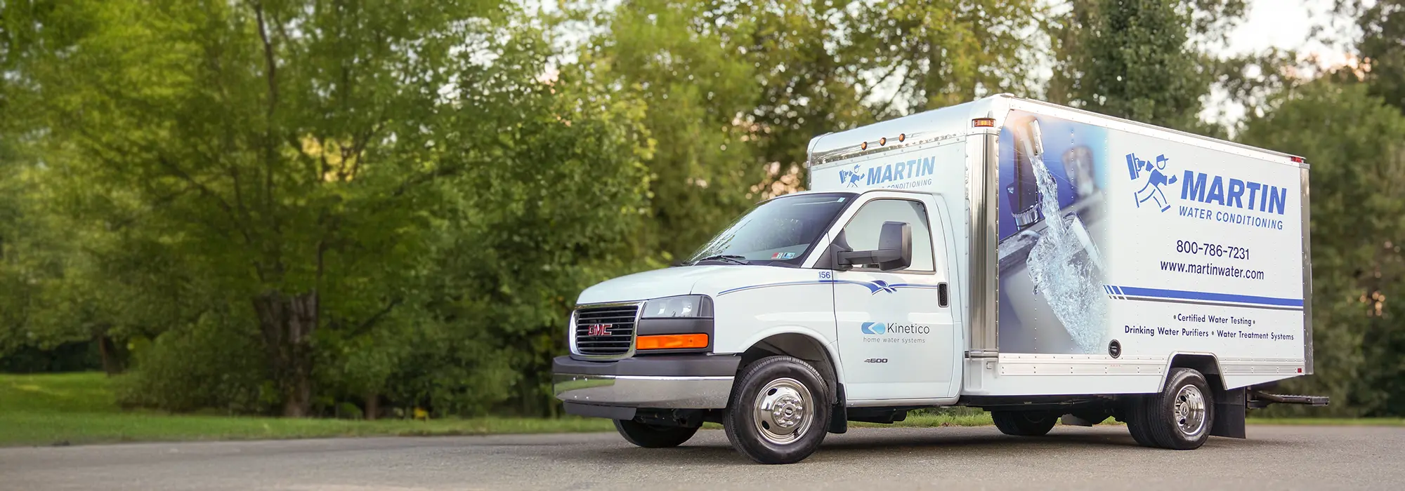 A white GMC box truck with Martin Water Conditioning branding is parked on a paved road, with trees and greenery in the background. The truck displays contact information and a large image of running water.