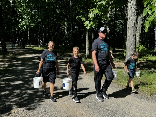Four people, including two children, walk on a sunlit forest path carrying white buckets. They wear matching black walk for clean water shirts. Trees and shade surround the group as they participate in an outdoor event.